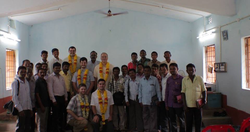 A large group photo in a classroom in India; several visitors wear marigold garlands